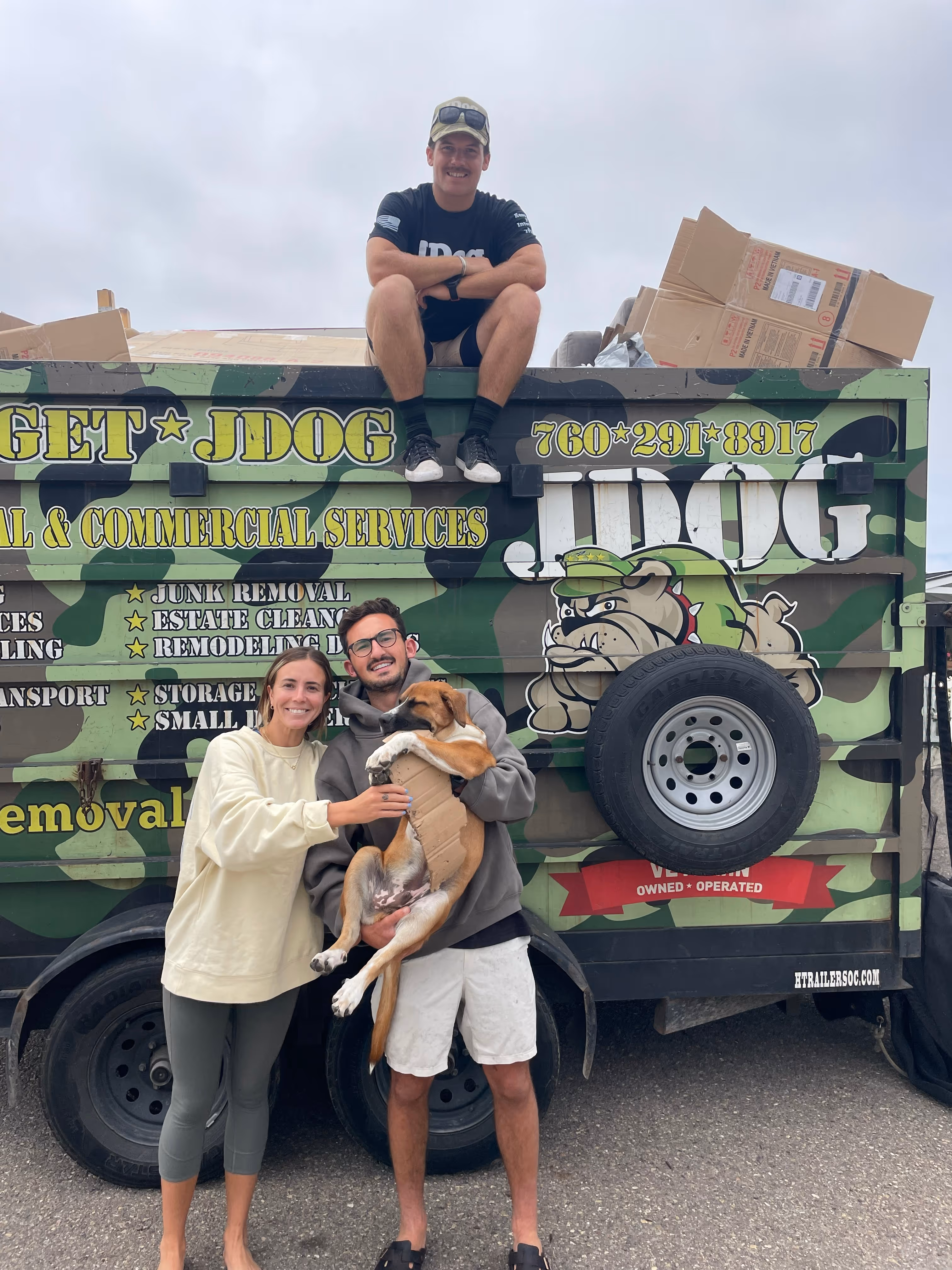 A couple holding their dog in front of the JDog trailer with a crew member sitting on top.